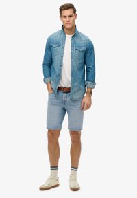Camisa de mezclilla de color azul claro, de mangas cortas, con dos bolsillos en el pecho. Camiseta blanca debajo. Pantalones cortos azules claros, zapatillas de deporte beiges y calcetines a rayas.