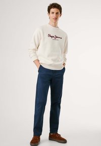Sudadera crema con logo rojo "Pepe Jeans London", combinada con pantalones azul marino y zapatillas marrones con cordones. La textura es suave; el ajuste es casual.