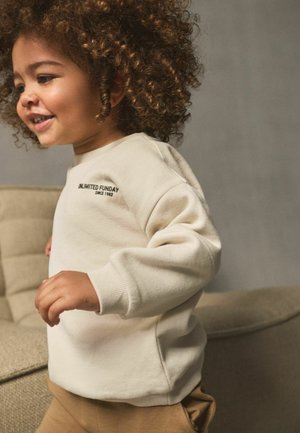 Sweatshirt crème avec des poignets côtelés arborant le texte "UNLIMITED FUNDAY". Texture douce, col rond, associé à un pantalon beige.