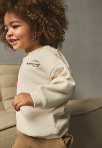 Krämfärgad sweatshirt med ribbade muddar som har texten "UNLIMITED FUNDAY". Mjuk textur, rund halsringning, ihopparad med beige byxor.