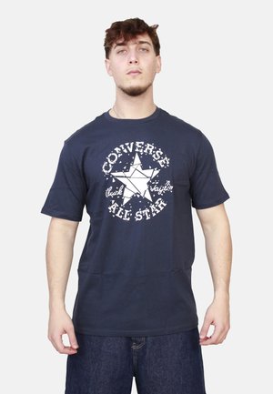 Giovane uomo che indossa una maglietta Converse All Star blu navy con un grande logo a stella e jeans blu scuro, in piedi davanti a uno sfondo uniforme.