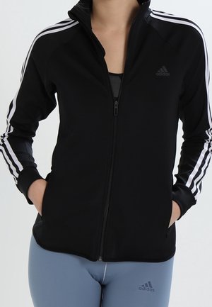 Zwarte Adidas ritsvest met witte strepen op de schouders, gedragen over een zwarte top, gecombineerd met lichtblauwe Adidas legging.