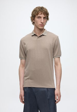 Ung mand med lyst brunt bølget hår iført en beige kortærmet polo med lodrette striber og mørkeblå bukser mod en ensfarvet baggrund.