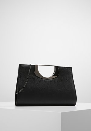 Sac à main noir texturé avec poignée métallique en demi-cercle et fine chaîne, posé sur un piédestal blanc sur fond gris uni.