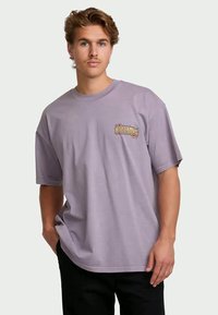 Jonge man met lichtbruin haar, gekleed in een loslavendel T-shirt met een kleine, kleurrijke grafiek op de borst en zwarte broek.