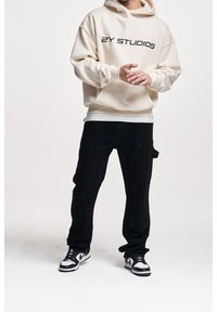 Kreemihuppari, jossa lukee mustalla "2Y STUDIOS", yhdistettynä mustiin farkkuihin. Jalan päällä musta-valkoisen sneaker-malliston kengät.