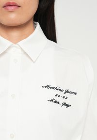 Balts krekliņš ar melniem izšūtiem vārdiem "Moschino Jeans 86.23 Milāna, Itālija" uz krūtīm, ko valkā cilvēks ar gariem tumšiem matiem.
