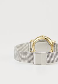 Relógio com bracelete de malha prateada e caixa em tom dourado, apresentando um mostrador redondo e um fecho de fecho, exibindo um design elegante e minimalista.