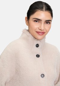 Jasnoróżowy, puszysty płaszcz z wysokim, falbaniastym kołnierzem. Posiada trzy czarne guziki z przodu. Miękka tekstura i luźny krój.