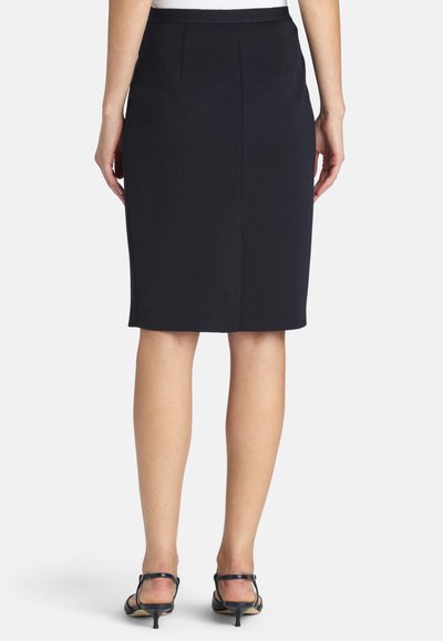 Kobieta w czarnej spódnicy ołówkowej do kolan i czarnych sandałach na niskim obcasie, pokazana od tyłu na jednolitym tle.