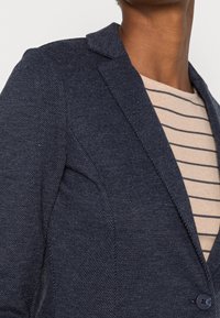 Tmavě modrý blazer s texturovaným herringbone vzorem, s jedním knoflíkem, s klopami, nošený přes světlý pruhovaný vršek.