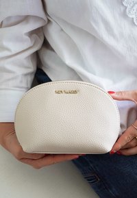 Pochette en cuir couleur crème avec une surface texturée, de forme arrondie, et le logo doré "HEY MARLY" à l'avant, tenue dans une main.