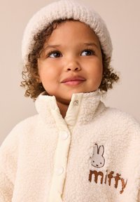 Kreemivärvi fliisjakke, millel on pehme tekstuur, varustatud nööbiga kinnitamisega ja tikandiga jänesemotiiviga, millel on tekst "miffy".