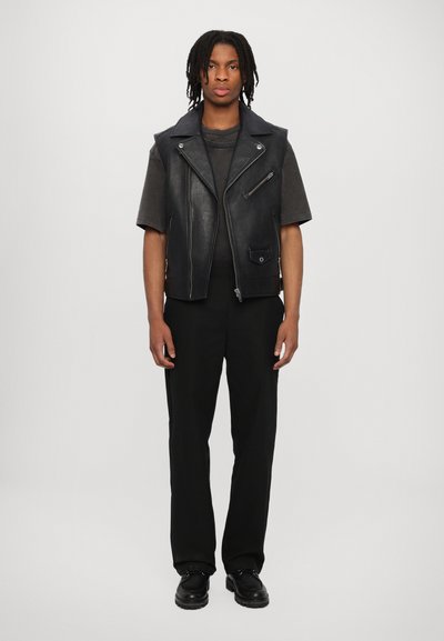 Zwart leren mouwloos vest met zilveren rits, gedragen over een donkergrijs T-shirt en gecombineerd met zwarte broeken en laarzen, staand tegen een neutrale achtergrond.