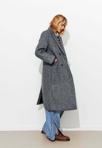 Manteau en laine gris à double boutonnage, avec poches latérales et col cranté. Associé à un jean à jambes larges et des chaussures à motifs.