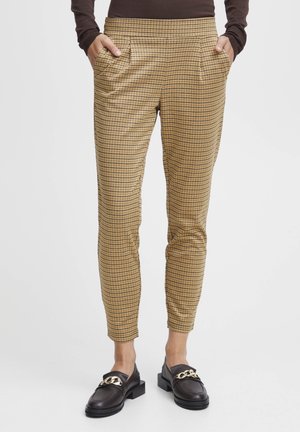 Personne portant un pantalon marron à carreaux fuselé, les mains dans les poches, et des mocassins noirs avec un détail de chaîne dorée.