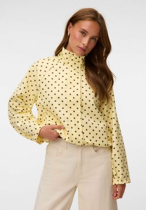 Femme portant un chemisier jaune à manches longues avec des pois noirs et un pantalon beige taille haute, posant avec une main dans la poche.