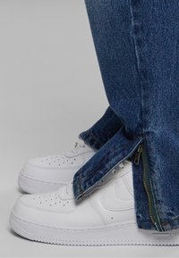 Witte sneakers met een geperforeerde neus en een gevoerde kraag, gecombineerd met donkerblauwe jeans met ritsdetails aan de zoom en een rechte snit.