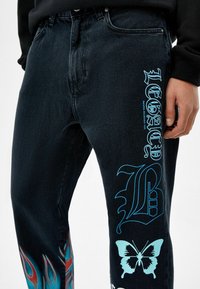 Jeans in denim nero con design grafici blu, inclusi testo e farfalla, con accenti di fiamma in rosso e turchese lungo il lato.