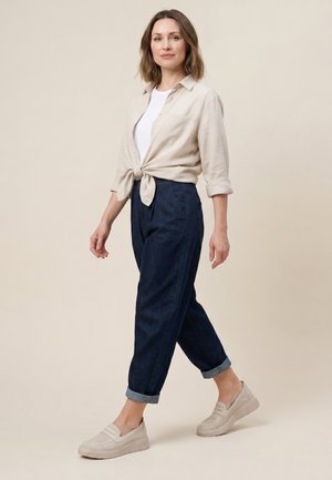 Kvinde, der går iført beige knyttet skjorte over hvid top, mørke opkrøllede bukser og beige slip-on sko mod ensfarvet baggrund.