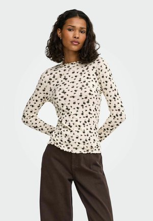 Junge Frau mit lockigem dunklem Haar, die ein cremefarbenes langärmliges Oberteil mit kleinem Blumenmuster und dunkelbraune Hose trägt, steht mit den Händen hinter dem Rücken.