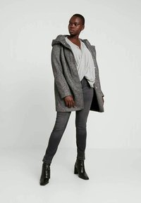 Casaco de mistura de lã cinza com capuz, apresentando um fecho de botão único, combinado com jeans skinny cinzas e botas de tornozelo pretas com tachas.