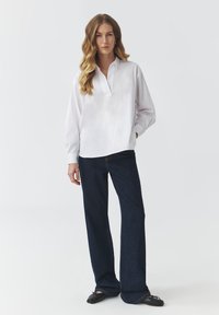 White long-sleeve shirt with a half-placket and relaxed fit, paired with dark blue flared jeans and black flats. Smooth, lightweight fabric.