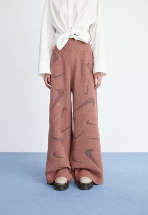 Pantalones marrones peludos de pierna ancha con patrones oscuros en forma de curva, combinados con camisa blanca anudada y botas beige gruesas sobre suelo azul.