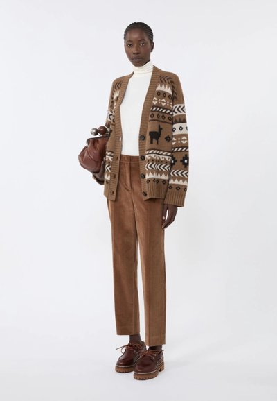 Cardigan marrone lavorato a maglia con motivi geometrici, dolcevita bianco, pantaloni in velluto a coste marroni e scarpe stringate marroni. Tiene una borsa marrone.