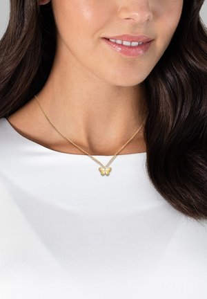 Collana con pendente a forma di farfalla in oro su una delicata catena, indossata sopra una camicia bianca. Il pendente presenta dettagli intricati e una finitura liscia.