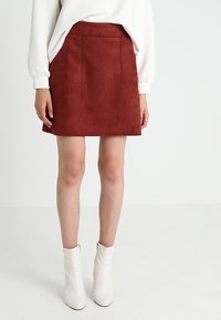 Jupe mini en suède couleur rouille avec deux poches avant, coupe droite et texture lisse, assortie à un pull blanc texturé et des bottines blanches.