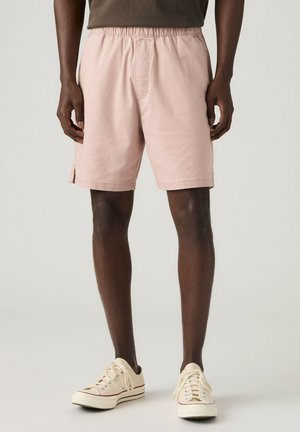 Man draagt lichtroze shorts met elastische taille en witte lage sneakers, staand tegen een effen achtergrond.