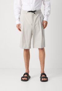 Pantaloni corti grigio chiaro su misura con cintura elastica e cordoncino nero. Indossati con sandali neri, dotati di una striscia e dettagli con fibbia.