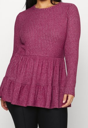 Femme portant un haut peplum magenta à manches longues en maille côtelée, associé à un pantalon noir, debout devant un fond blanc.