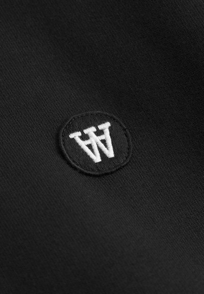 Tissu noir avec un logo brodé circulaire comportant les lettres blanches "W" et "A". La texture est lisse avec un léger aspect satiné.