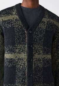 Temno zelena in črna jopica s vzorcem, z V-izrezom, teksturiranim pletenjem, šestimi črnimi gumbi in kontrastnimi barvnimi poudarki.