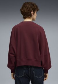 Bordeaux bomulds sweatshirt med afslappet pasform, sænkede skuldre og brede manchetter, parret med mørke denim jeans. Enkel, ribbet detalje ved halsen.
