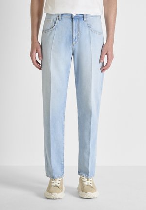 Lichtblauwe jeans met rechte pijpen, gecombineerd met beige sneakers en een wit overhemd dat in de broek is gestopt, zichtbaar van taille tot schoenen.