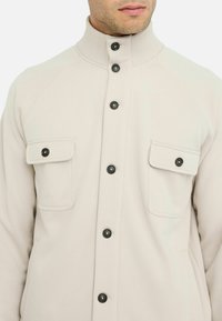 Giacca beige con bottoni, colletto alto, bottoni neri e due tasche sul petto. Presenta una texture liscia e maniche raglan.