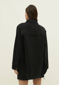 Chaqueta de denim negra de gran tamaño con cuello, puños abotonados y dobladillo sin terminar. Presenta una textura descolorada y un detalle de correa en la parte trasera.