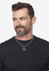 Silverfärgad kedjehalsband med en fredssymbolhängsmycke, bärs över en enkel svart t-shirt; minimalistisk design, slät textur.