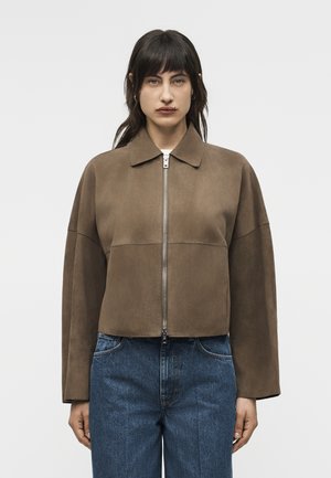 Egyenes, sötét hajú nő barna, zipzáros szarvasbőr dzsekit és kék farmert viselve, sima fehér háttér előtt áll.