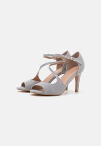 Silver high-heeled sandals with a textured fabric upper, peep-toe design, and crossed ankle straps, featuring a smooth, light-colored insole.