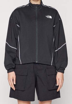 Persoon draagt een zwart jack met ritssluiting en bijpassende cargoshorts met elastische boorden, voorzien van witte reflecterende biezen en het logo van The North Face.