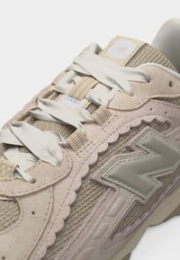 Κοντινό πλάνο μπεζ αθλητικού παπουτσιού New Balance που δείχνει υφές από δίχτυ, σουέτ, φαρδιές σατινένιες κορδόνια και το αντανακλαστικό λογότυπο "N" της μάρκας στο πλάι.