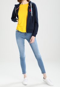 Námořnická mikina na zip s bílými tkaničkami, vrstvená přes žluté tričko, spárovaná se světle modrými skinny džínami a bílými teniskami.