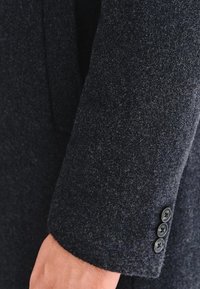 Mroczny szary wełniany rękaw płaszcza o fakturowanym wykończeniu, z trzema czarnymi guzikami przy mankiecie i dopasowanym krojem.