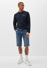 Marineblaues Langarmhemd mit Logo, Jeanskurze Hosen und weiße High-Top-Sneaker. Der Denim ist leicht ausgebleicht mit einer lässigen Passform und einem umgeschlagenen Saum.