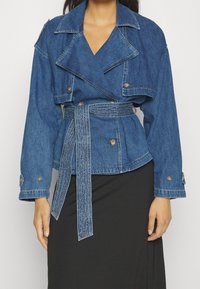 Denimjacka med åtdragen midja, dubbelknäppning fram, breda slag och långa ärmar. Har en knytbälte och knappringar.