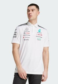 White polo shirt with collar, featuring black accents and multiple logos on the front, made of a smooth, slightly textured fabric.